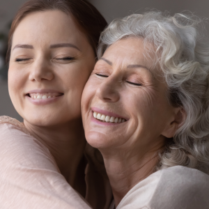 A white grandmother with white hair hugging her granddaughter while closing her eyes. Her granddaughter has auburn hair and is also smiling with her eyes closed.