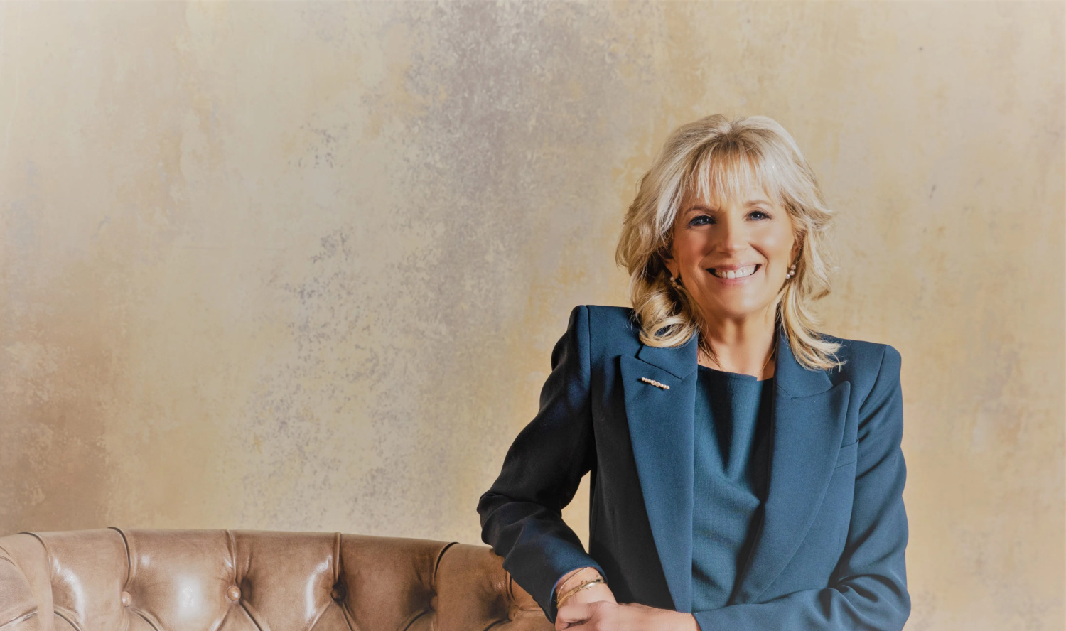 First Lady Dr. Jill Biden has shoulder-length blonde hair and smiles at the camera. She is seated on the arm of a brown leather sofa and wearing a blue suit with pearl earrings, a pearl brooch on her right lapel, and gold bracelets on her right wrist.