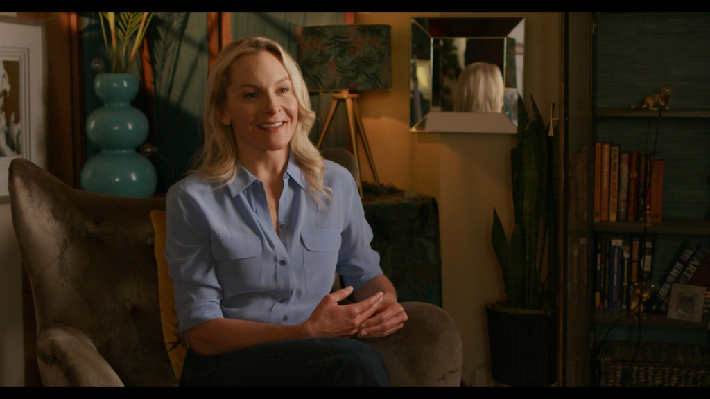 Joslyn DeFreece, a white woman with long blonde hair and wearing a blue button-down shirt, is seated and smiling while talking on the set of the short film "Pieces of Me."