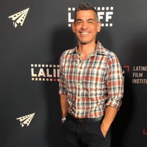 An image of Nick Oceano Armendariz, a young Latino man wearing a checked shirt and dark pants