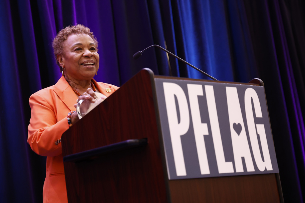 Rep. Barbara Lee, wearing an orange suit, holds a heart-shaped glass award and smiles while speaking at a podium. PFLAG's logo is on the front of the wood podium.