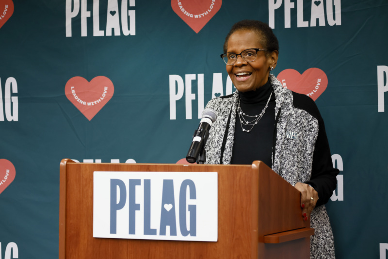 Edith Guffey, who wears cat-eye glasses, earrings, and a multi-strand necklace over a high-necked blouse and sweater jacket, stands at a wooden podium. Behind her is a backdrop with the PFLAG logo and hearts that read "leading with love."