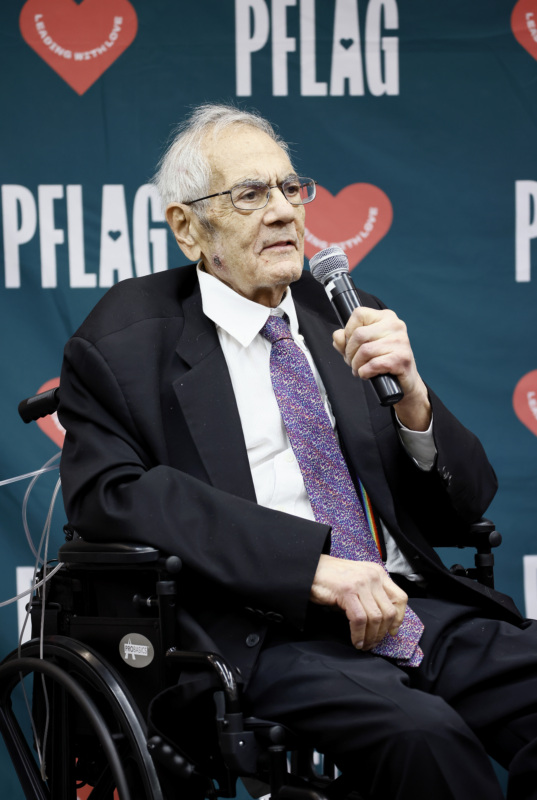 Retired Rep. Barney Frank, who wears glasses and a suit and tie, is seated in a wheelchair. Behind him is a backdrop patterned with the PFLAG logo and hearts that read "leading with love."