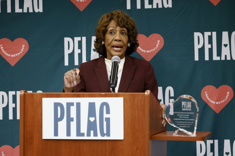 Rep. Maxine Waters, who wears large pearl earrings and a blazer over a silk top, is standing at a wooden podium. Behind her is a backdrop with the PFLAG logo and hearts that read, "leading with love." To the right of the podium is the engraved heart-shaped crystal award for the PFLAG Champion of Justice.