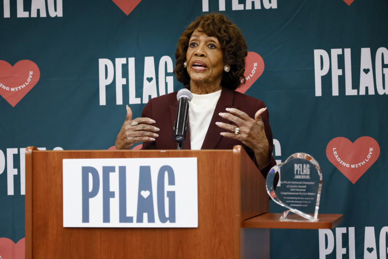 Rep. Maxine Waters, who wears large pearl earrings and a blazer over a silk top, is standing at a wooden podium. Behind her is a backdrop with the PFLAG logo and hearts that read, "leading with love." To the right of the podium is the engraved heart-shaped crystal award for the PFLAG Champion of Justice.