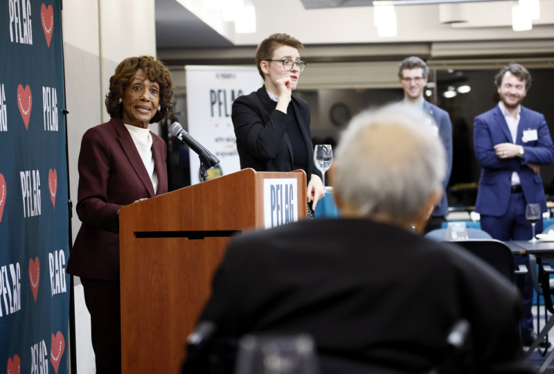 Retired Rep. Barney Frank is seated and faces away from the camera toward Rep. Maxine Waters as she speaks at the podium. Next to her is a sign-language interpreter.