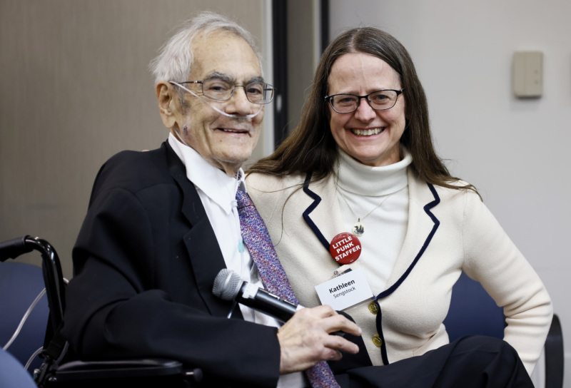 Retired Rep. Barney Frank, who wears glasses, a suit and tie, sits next to Kathleen Sengstock, who wears a big button that reads "Little Punk Staffer."