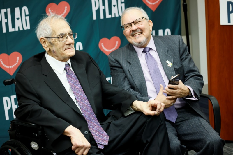 Retired Rep. Barney Frank, who wears glasses and a suit and tie, and Diego Sanchez, wearing glasses, a suit and tie, and holding a cell phone, sit together and laugh.