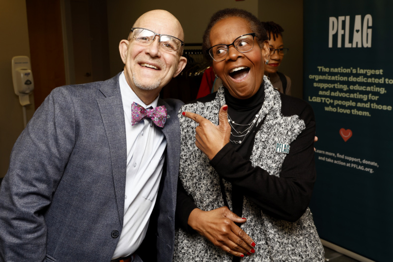 Brian Bond, who wears glasses and a suit with a bowtie, leans in with a smile toward Edith Guffey, who also has a big smile and laughing eyes as she points to Brian. Edith is wearing cat-eye glasses, earrings, a multi-string necklace, and a sweater jacket.