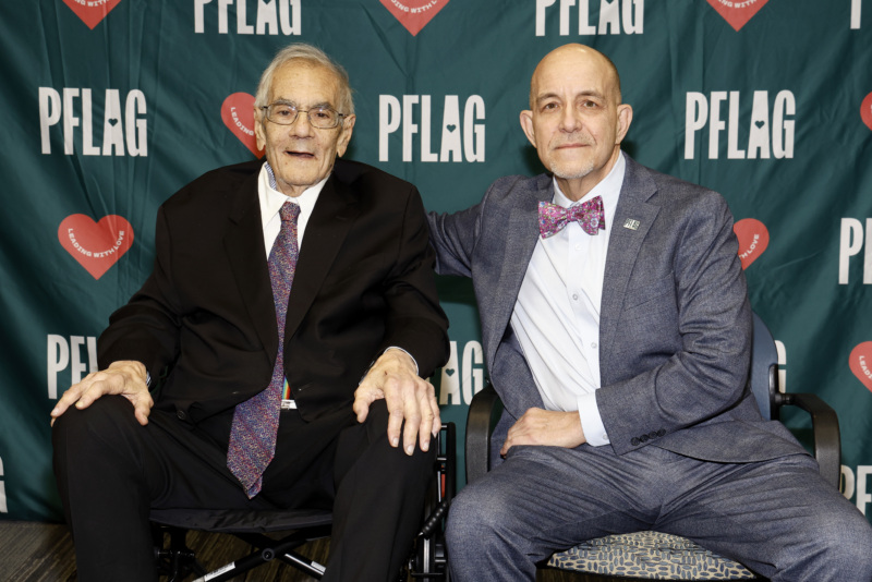 Retired Rep. Barney Frank, who wears glasses and a suit and tie, is seated in a wheelchair to the left of PFLAG National CEO Brian Bond, who wears a suit and bowtie and has his arm around Rep. Frank. Both are seated in front of a backdrop patterned with the PFLAG logo and hearts with the words "leading with love."