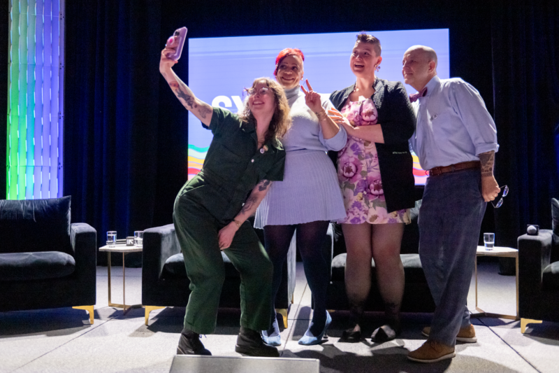 Casey McQuiston, who wears green coveralls holds up a phone to take a selfie that includes a smiling Nicole Hannah-Jones, who wears a lavender sweater dress and leggings and flashes a peace sign; Sam Helmick, who wears a flower print dress with a black cardigan, and Brian K. Bond, who wears a bow tie and dress shirt with the sleeves rolled up.