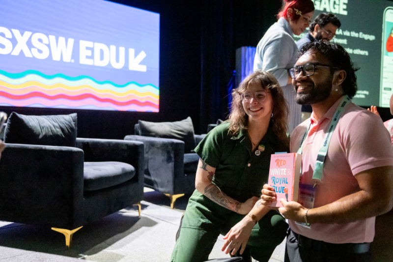 Author Casey McQuiston wears green coveralls and poses with a fan who has a beard and glasses while holding a copy of their banned book "Red, White and Royal Blue". They are standing on the stage at SXSW EDU and are looking toward another photographer who is not pictured.